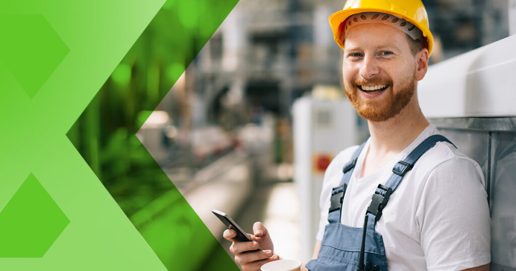 Happy temporary worker in factory, wearing a hard yellow hat. He is smailing at the camera and is holding a mobile phone