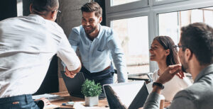 Staffing professional shaking hadns with client, leaning over a table in an office, with colleagues looking on, everyone smiling.